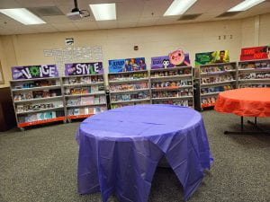 This is a picture of Scholastic carts with books on each shelf. Each cart has a sign above it advertising books. There are tables covered with a tablecloth in front of the carts for more book displays.
